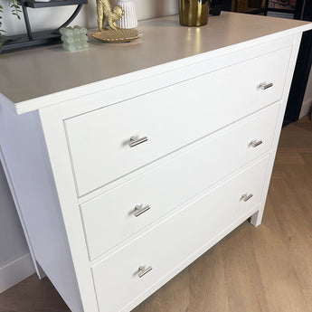 Een witte commode met drie lades en Remo RVS knoppen van yessential.nl staat op een lichte houten vloer. Een gouden apenbeeldje en een groen kralenornament sieren de bovenkant van de commode.