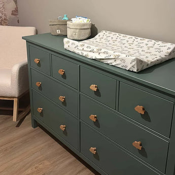 De Bambino Cloud commode van yessential.nl in groen, met negen lades en houten meubelknop, heeft een aankleedkussen met bloemmotief en manden voor babyspullen - ideaal voor de kinderkamer naast een beige glijder op een houten vloer.