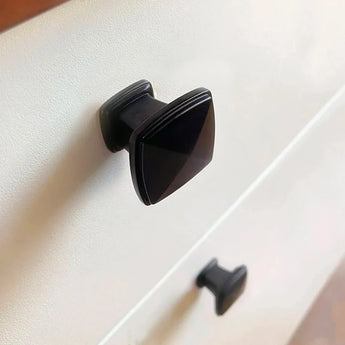 Close-up van een witte lade met twee verticaal uitgelijnde Adaro Black Knobs van yessential.nl. De vierkante handgrepen zijn gemonteerd op een glad, vlak oppervlak, boven elkaar.