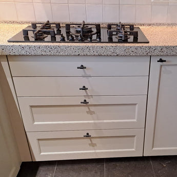 Een gaskookplaat met vier branders is geplaatst in een gespikkeld aanrechtblad boven lades en een kast, elk voorzien van Cordero Black Knobs van yessential.nl. De keuken is afgewerkt met een wit betegelde achterwand.