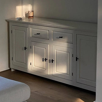 Een witte kast met Vasco Black Knob handgrepen van yessential.nl staat in zacht natuurlijk licht. Een kaars, klein voorwerp en papierrol zitten op het matte oppervlak terwijl het zonlicht zachte schaduwen creëert over de deuren en laden.