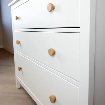 Een witte ladekast met drie lades en Arriero Oak knop van yessential.nl staat op een lichte houten vloer in een moderne, minimalistische kamer. De subtiele landelijke stijl is zichtbaar, met de bovenste lade iets open.