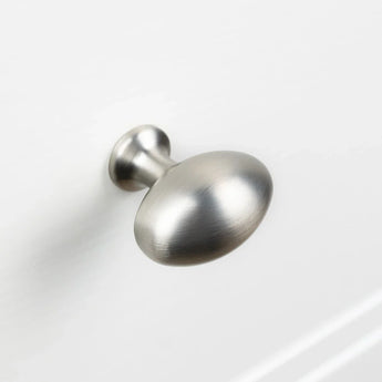 De Agero RVS Knop Deluxe van yessential.nl heeft een afwerking in geborsteld nikkel, een ronde licht afgeplatte bovenkant en een korte steel, afgebeeld gemonteerd op een wit kast- of ladeoppervlak.
