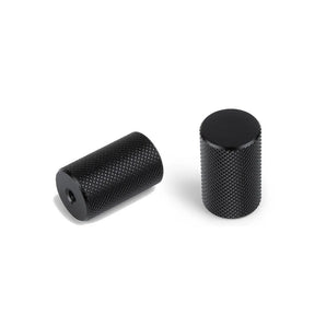Twee zwarte cilindervormige metalen knoppen met gestructureerd oppervlak van yessential.nl, waaronder de Remo Black Deluxe Knob, worden getoond op een witte achtergrond. De ene knop staat rechtop, terwijl de andere op zijn kant ligt en een gat in het midden laat zien.