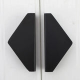 Twee zwarte, geometrische, ruitvormige boekensteunen van yessential.nl, genaamd Triano Black 64mm, staan rechtop op een wit houten oppervlak met schuine randen naar buiten en rechte randen die in het midden samenkomen.