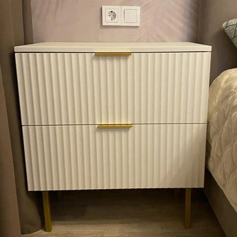 Het Enzo Gold nachtkastje van yessential.nl, met verticale groeven, messing handgrepen en gouden poten, staat bij het bed. Erboven aan de muur zitten een stopcontact en een lichtschakelaar.
