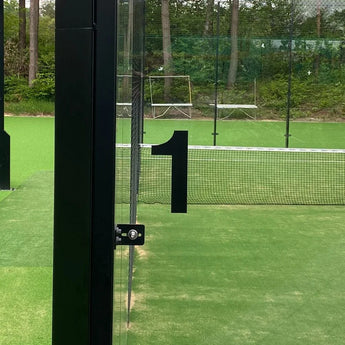Een glazen deur met een groot Animo Black 15cm huisnummer "1" van yessential.nl (geen boormachine) opent naar een groene buiten paddle tennisbaan met net, omgeven door bomen en gaashekwerk.