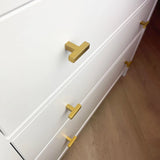 Een close-up van een witte commode met Vigio Gold Knobs van yessential.nl op drie lades, elk versierd met moderne gouden T-vormige handgrepen, bovenop een lichtbruine houten vloer.
