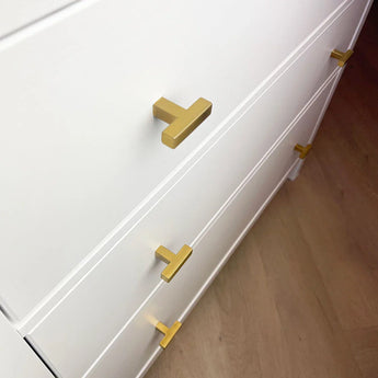 Een close-up van een witte commode met Vigio Gold Knobs van yessential.nl op drie lades, elk versierd met moderne gouden T-vormige handgrepen, bovenop een lichtbruine houten vloer.