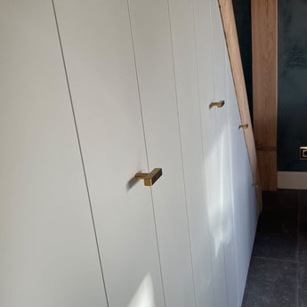 Een rij witte inbouwkasten met luxe gouden afwerking en Vasco Gold Knob van yessential.nl is geïnstalleerd onder een houten trap, naast een donkerblauwe muur en grijze tegelvloer.