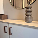 Het Vasco Walnut dressoir van yessential.nl, met walnotenhouten handgrepen, staat onder een ronde wandspiegel. Een bruin gestructureerde vaas met gedroogde takken siert de bovenkant, terwijl een rieten mand aan de linkerkant staat.