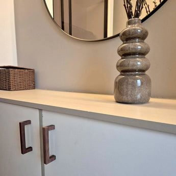 Het Vasco Walnut dressoir van yessential.nl, met walnotenhouten handgrepen, staat onder een ronde wandspiegel. Een bruin gestructureerde vaas met gedroogde takken siert de bovenkant, terwijl een rieten mand aan de linkerkant staat.