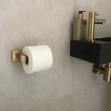 De Vero Gold Deluxe toiletrolhouder van yessential.nl is gemonteerd op een betegelde muur naast een moderne zwarte wastafel met messing armaturen en een zeepdispenser, waardoor luxe badkameraccessoires in de ruimte komen.