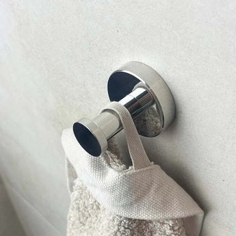 De Mano Chroom Wc-set van yessential.nl heeft een slank chromen wandhaakje dat een beige handdoek vasthoudt aan de lus, wat stijl toevoegt aan je badkameraccessoires tegen een lichtgekleurde muur.