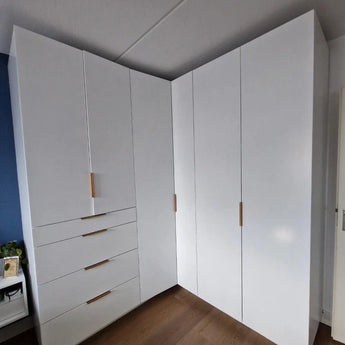 De Enzo Rose Gold hoekkast van yessential.nl heeft glanzend witte deuren, moderne handgrepen, vier lades aan de linkerkant, hoge kasten aan beide kanten en staat elegant op een houten vloer in een moderne kamer.
