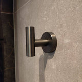 De Rodrigo Gunmetal Haak van yessential.nl heeft een slank, cilindervormig metalen ontwerp. Gemonteerd op een grijze betegelde muur, voegt het een moderne touch en subtiele schaduw toe - ideaal voor elke badkamer.
