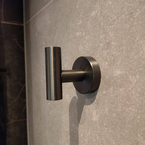 De Rodrigo Gunmetal Haak van yessential.nl heeft een slank, cilindervormig metalen ontwerp. Gemonteerd op een grijze betegelde muur, voegt het een moderne touch en subtiele schaduw toe - ideaal voor elke badkamer.