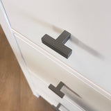 Close-up van een wit ladekastje met Vigio Gunmetal Knob handgrepen van yessential.nl, van bovenaf gezien onder een hoek. De moderne T-vormige knoppen accentueren het gladde oppervlak en zijn een voorbeeld van hedendaags design. De vloer verschijnt op de achtergrond.