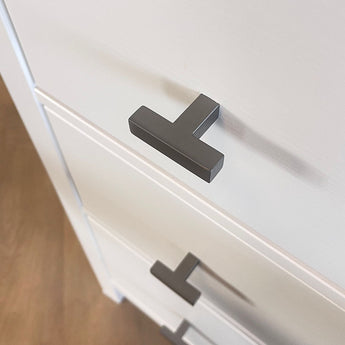 Close-up van een wit ladekastje met Vigio Gunmetal Knob handgrepen van yessential.nl, van bovenaf gezien onder een hoek. De moderne T-vormige knoppen accentueren het gladde oppervlak en zijn een voorbeeld van hedendaags design. De vloer verschijnt op de achtergrond.