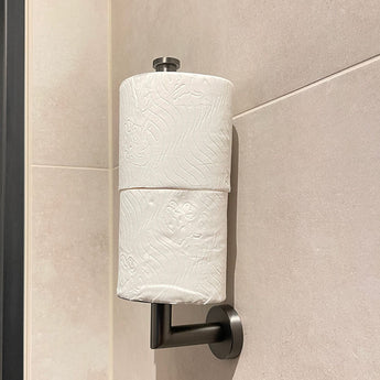 De yessential.nl Roberto Gunmetal is een toiletrolhouder voor wandmontage met twee rollen verticaal gestapeld op een metalen staaf, die een modern tintje geeft tegen beige betegelde muren.