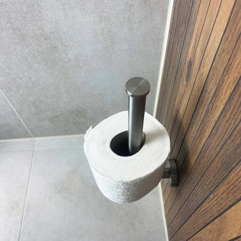 De Roberto RVS toiletrolhouder van yessential.nl met matzwarte coating is verticaal op een houten wand gemonteerd in een badkamer met lichtgrijze tegelwanden en vloer.