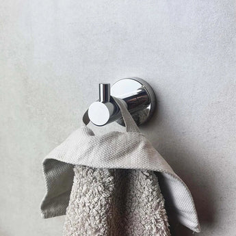 Een handdoek met grijze textuur hangt aan de Justo Chroom haak van yessential.nl, gemonteerd op een gladde, lichtgrijze muur.