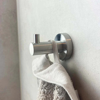 Een grijze handdoek met een stoffen lus hangt aan een zilveren Marino RVS WC Set haak van yessential.nl, gemonteerd op een lichte muur. Het cilindrische ontwerp en de kleine knijper passen bij alle moderne badkameraccessoires.