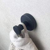 Een matzwarte wandhaak uit de yessential.nl Midao Black WC set is gemonteerd op een lichte muur en houdt een beige handdoek vast aan de stoffen lus.