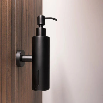 Het Alberto Black zeeppompje van yessential.nl is aan de muur bevestigd op een houten paneel, waardoor het strakke moderne ontwerp wordt benadrukt. De rechterkant van het beeld toont een effen, lichtgetinte achtergrond.