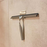 De Zamoro RVS van yessential.nl, een roestvrijstalen douche trekker met zwarte rubberen blad, hangt aan een muurhaak met 3M ophangsysteem op beige tegels.