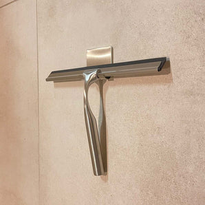 De Zamoro RVS van yessential.nl, een roestvrijstalen douche trekker met zwarte rubberen blad, hangt aan een muurhaak met 3M ophangsysteem op beige tegels.