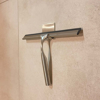 De Zamoro RVS van yessential.nl, een roestvrijstalen douche trekker met zwarte rubberen blad, hangt aan een muurhaak met 3M ophangsysteem op beige tegels.