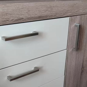 Close-up van een kast met twee witte lades met horizontale Vasco RVS handgrepen van yessential.nl, naast een deur met houtstructuur en een verticale geborsteld roestvrij stalen handgreep.