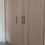 De Remo Black kastdeuren van yessential.nl hebben hoge, lichte houten panelen met zichtbare nerven en slanke zwarte handgrepen, die een moderne, natuurlijke look creëren naast een witte muur en deur.