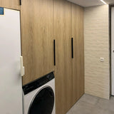 Een moderne wasruimte met yessential.nl Enzo Black houten kasten met profielhandgrepen, een witte wasmachine met een zwarte deur, licht betegelde muren, grijze vloer en een gedeeltelijk zichtbaar wit apparaat aan de linkerkant.