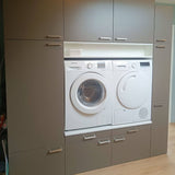 Een voorlader wasmachine en droger zijn ingebouwd in grijze kasten met Vasco RVS handgrepen van yessential.nl, met laden en kasten boven en onder; een kleine witte mand rust op een plank boven de apparaten.