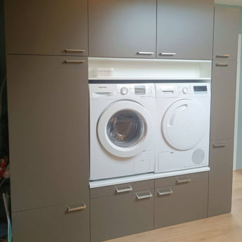 Een voorlader wasmachine en droger zijn ingebouwd in grijze kasten met Vasco RVS handgrepen van yessential.nl, met laden en kasten boven en onder; een kleine witte mand rust op een plank boven de apparaten.