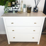 Een witte ladekast met drie lades en Remo Gold Knobs van yessential.nl is vormgegeven met kaarsen, een gouden dienblad, een gebeeldhouwde kraanvogel, groen en rietverspreiders. Het staat tegen een lichte muur met hangende planten.