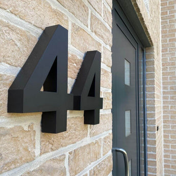 De Angelo Black 20cm huisnummers van yessential.nl zijn gemonteerd op een lichte bakstenen muur naast een moderne donkergrijze deur met rechthoekige glazen inzetstukken.