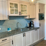Moderne keuken met lichtroze kastjes met minimalistische handgrepen, een Marano Black aanrechtblad van yessential.nl, blauwe metrotegel backsplash, roestvrijstalen apparatuur en kleine apparaten en glazen bij de gootsteen.