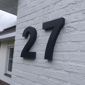 Het Angelo Black 30cm huisnummer van yessential.nl is als opvallend groot zwart cijfer op een witte bakstenen muur gemonteerd, met een raam en dak op de achtergrond die het moderne design onderstrepen.
