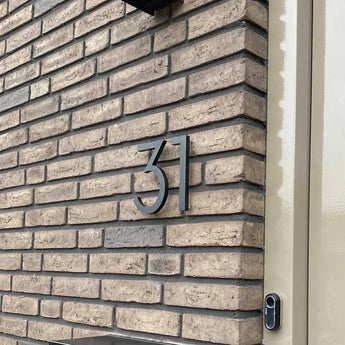 Het Milano Black 15cm huisnummer van yessential.nl is gemonteerd op een beige bakstenen muur bij een lichtgekleurde deur, met een deurbel gedeeltelijk zichtbaar aan de rechterrand.