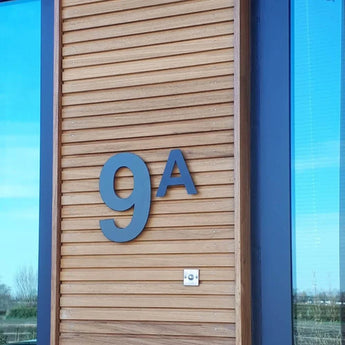 Het Adamo Black 30cm RVS huisnummer van yessential.nl siert een houten gevel naast een raam. Onder het zwevende zwarte huisnummer bevindt zich een kleine zilveren intercom of deurbel. In het glas weerspiegelen bomen en lucht.