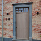 Een lichte houten voordeur met rechthoekige ramen erboven, gevat in een grijze stenen omlijsting. De lichtrode bakstenen muur is voorzien van een yessential.nl Adamo Black 15cm huisnummer "51B" aan de linkerkant en twee zwarte wandlampen flankeren de deur.