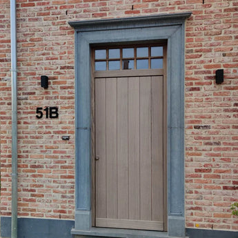 Een lichte houten voordeur met rechthoekige ramen erboven, gevat in een grijze stenen omlijsting. De lichtrode bakstenen muur is voorzien van een yessential.nl Adamo Black 15cm huisnummer "51B" aan de linkerkant en twee zwarte wandlampen flankeren de deur.