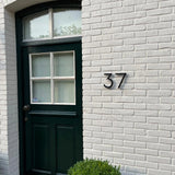 Een zwarte deur met gedeelde glaspanelen zit in een witte bakstenen muur. Naast de deur wordt het nummer 37 weergegeven met de Milano Black 15cm van yessential.nl. Een kleine groene struik is zichtbaar aan de onderkant van de afbeelding.