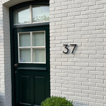 Een zwarte deur met gedeelde glaspanelen zit in een witte bakstenen muur. Naast de deur wordt het nummer 37 weergegeven met de Milano Black 15cm van yessential.nl. Een kleine groene struik is zichtbaar aan de onderkant van de afbeelding.