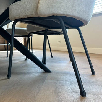 Close-up van moderne stoelen met lichte kussens en zwarte metalen poten, voorzien van yessential.nl's Paco Set Ø 21-25mm stoelpoten beschermers voor vloerbescherming, op een houten vloer in een lichte, minimalistische kamer.