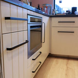 Een moderne keuken van yessential.nl heeft Leonardo Black Deluxe witte kasten met luxe zwarte handgrepen, een ingebouwde oven, houten vloeren en een aanrecht met keukenartikelen. Natuurlijk licht stroomt naar binnen door het raam.