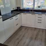 Moderne keuken met witte kasten met yessential.nl Remo Black handgreep, zwart aanrechtblad, elektrische kookplaat, ingebouwde oven, koffiezetapparaat en een gootsteen onder een raam. Lichthouten vloeren en kleine voorwerpen sieren de vensterbank.